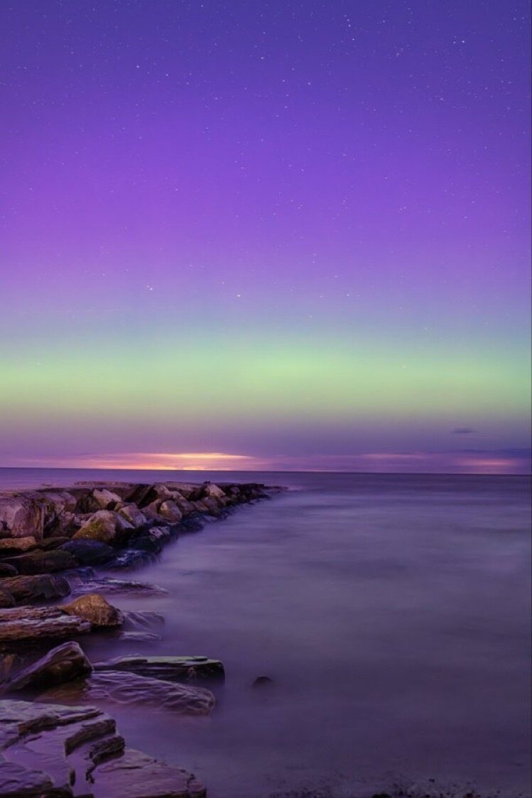 Aurora over a rocky breakwater at twilight — green and lavender bands above still water.
