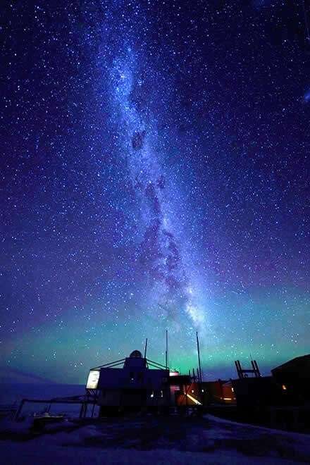 The Milky Way rising over a small lit outpost, with green airglow low on the horizon.